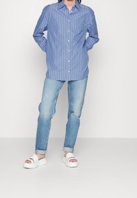 Blau-weiß gestreiftes Hemd mit Knöpfen, hellblaue Jeans und weiße Slides-Sandalen. Das Hemd hat einen klassischen Kragen und eine lässige Passform.