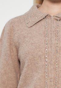 Strikkegenser i myk rosa med ribbestrikket krage, knappelukking foran og pyntet med sølv rhinestones langs knappeklaffen og kragen.