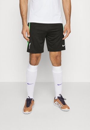 Mann trägt schwarze Liverpool-Fußballshorts mit grünen Streifen, weiße Socken mit lila Nike-Swoosh und kupferfarbene Nike-Fußballschuhe mit schwarzen Schnürsenkeln.