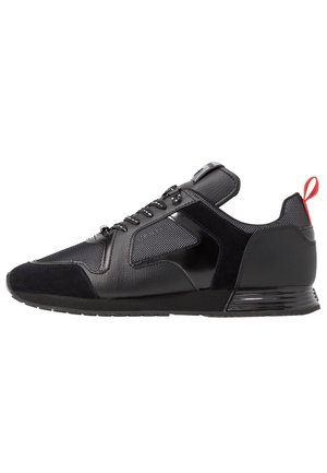 Sort lavtop sneaker med blandede læder- og meshpaneler, sorte snørebånd og en rød trækflap på hælen, vist fra siden på en hvid baggrund.