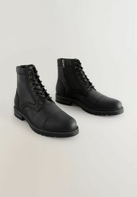 Botas de tobillo de cuero negro con cordones en la parte delantera, cuello acolchado y cremallera lateral. Suela de goma gruesa con diseño de puntera cap.