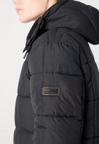 Black puffer jacket with a textured surface, featuring a hood, defined stitching, and a branded patch on the left shoulder.