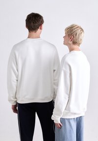 Weiße Sweatshirts mit Rundhalsausschnitt an zwei Personen, ausgestattet mit gerippten Bündchen und Saum, lässiger Passform und weicher Textur. Eines kombiniert mit dunklen Hosen, das andere mit heller Denim.