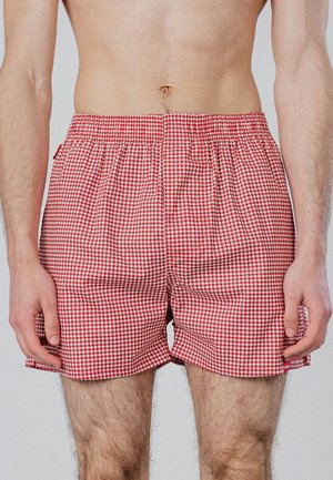 Männlicher Torso, der rote und weiße Badeshorts mit Hahnentrittmuster und elastischem Bund vor einfarbigem Hintergrund trägt.
