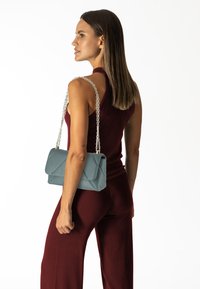 Borsa a tracolla in pelle verde acqua con un design trapuntato, tracolla in catena argentata e chiusura a flap, abbinata a un top senza maniche bordeaux e pantaloni.