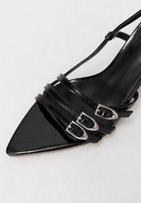 Sandale à talon haut noire à bout pointu avec trois fines lanières et boucles argentées sur le pied.