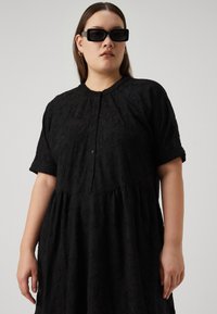 Μαύρο φόρεμα με υφή ανθοστοιχίας, κοντά μανίκια, κουμπιά μπροστά και σχέδιο φούστας χαλαρής, με πτυχίωση. Διακοσμημένο με γυαλιά ηλίου.