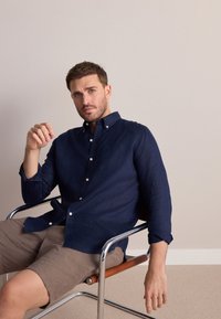 Camicia di lino blu navy con design con bottoni, colletto appuntito e taschino singolo sul petto; indossata con pantaloncini beige, seduto su una sedia cromata.