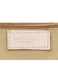 Kreemikas nahast silt, mis on õmmeldud beeži kanga tagakülge, kannab kirjet "Gusti Leder MADE IN ITALY" pruuni tõmbluku all.