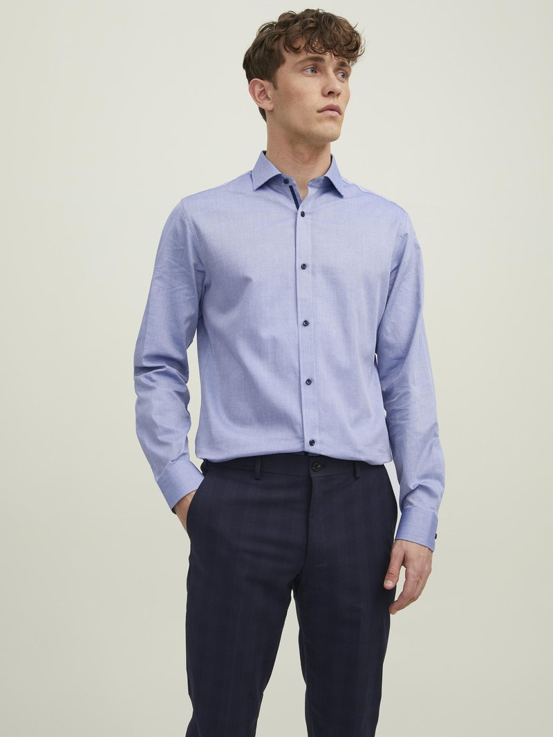 Camicia a maniche lunghe a quadri blu chiaro con colletto button-down. Accenti blu sulla patta e bottoni scuri. Abbinata a pantaloni a quadri scuri.