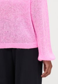 Rosa oversized stickad tröja med en lös, texturerad väv och klockärmar, tillsammans med svarta byxor.