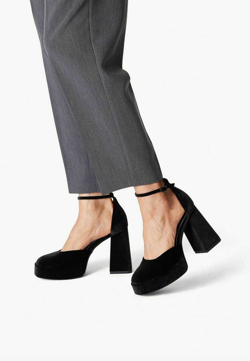 Personne portant un pantalon gris et des chaussures à plateforme à talons hauts en velours noir avec des brides à la cheville, debout devant un fond blanc.