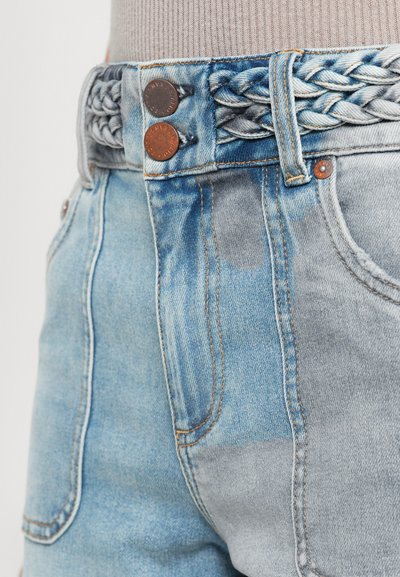 Gros plan sur un jean en denim patchwork bleu clair avec une ceinture tressée et deux boutons en métal à la taille.