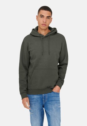 Jeune homme aux cheveux courts portant un sweat à capuche vert foncé et un jean bleu, debout avec une main dans la poche sur fond blanc.