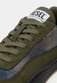 Sneaker in suede verde con sovrapposizione testurizzata, lacci piatti e etichetta con logo. Presenta un motivo multicolore sul lato e suola nera.