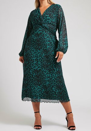 Kobieta ubrana w midi sukienkę w turkusowo-czarny wzór w cętki, z długimi rękawami i dekoltem w serek, oraz czarne szpilki z paskiem wokół kostki na jednolitym tle.