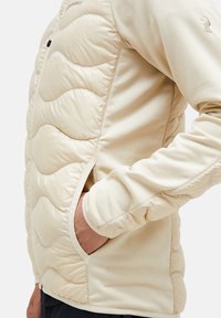 Beige hybrid jakke med en kviltet front med bølge mønstre, strækstofærmer og sidelommer. Blød tekstur med et strømlinet design.