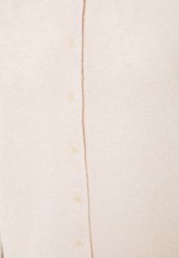 Gros plan sur un cardigan en maille beige avec une rangée de petits boutons assortis fermés au centre avant.