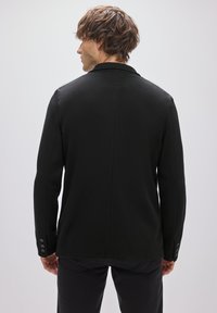 Blazer negro hecho de tela texturizada, con un corte entallado, cuello con solapas y adornos con botones en los puños. La vista trasera muestra un diseño sin costuras.