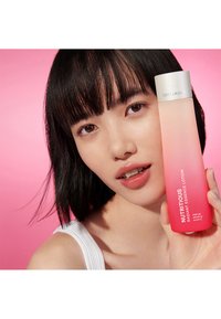 Estée Lauder Nutritious Radiant Essence Lotion i en gradient rosa flaske. Flasken har hvit tekst og en sølvhett. Bakgrunnen er rosa.