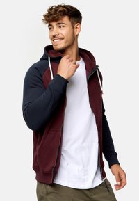 Sudadera con cremallera en color burdeos y azul marino, con cordón, mangas raglán, bolsillo canguro y texturas en contraste. Lleva puesta una camiseta blanca debajo.