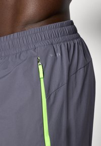 Grå atletiske shorts med elastisk talje, med en sidelomme med lynlås i en lys grøn farve og "WINCEL" branding.