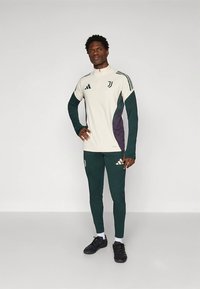 Športová tepláková súprava v krémovej, tmavozelenej a fialovej farbe. Má vysoký golier, dlhé rukávy a úzke nohavice s logom.