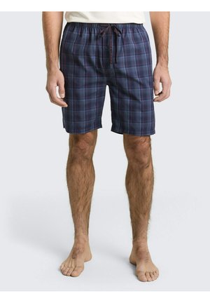 Mann trägt marineblaue und dunkelrote kariertem Shorts mit Kordelzug, steht barfuß vor einem einfarbigen hellen Hintergrund.