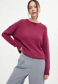Maglione fucsia lavorato a maglia con maniche lunghe e scollatura rotonda, abbinato a pantaloni grigi chiari su misura. La trama appare morbida e accogliente.