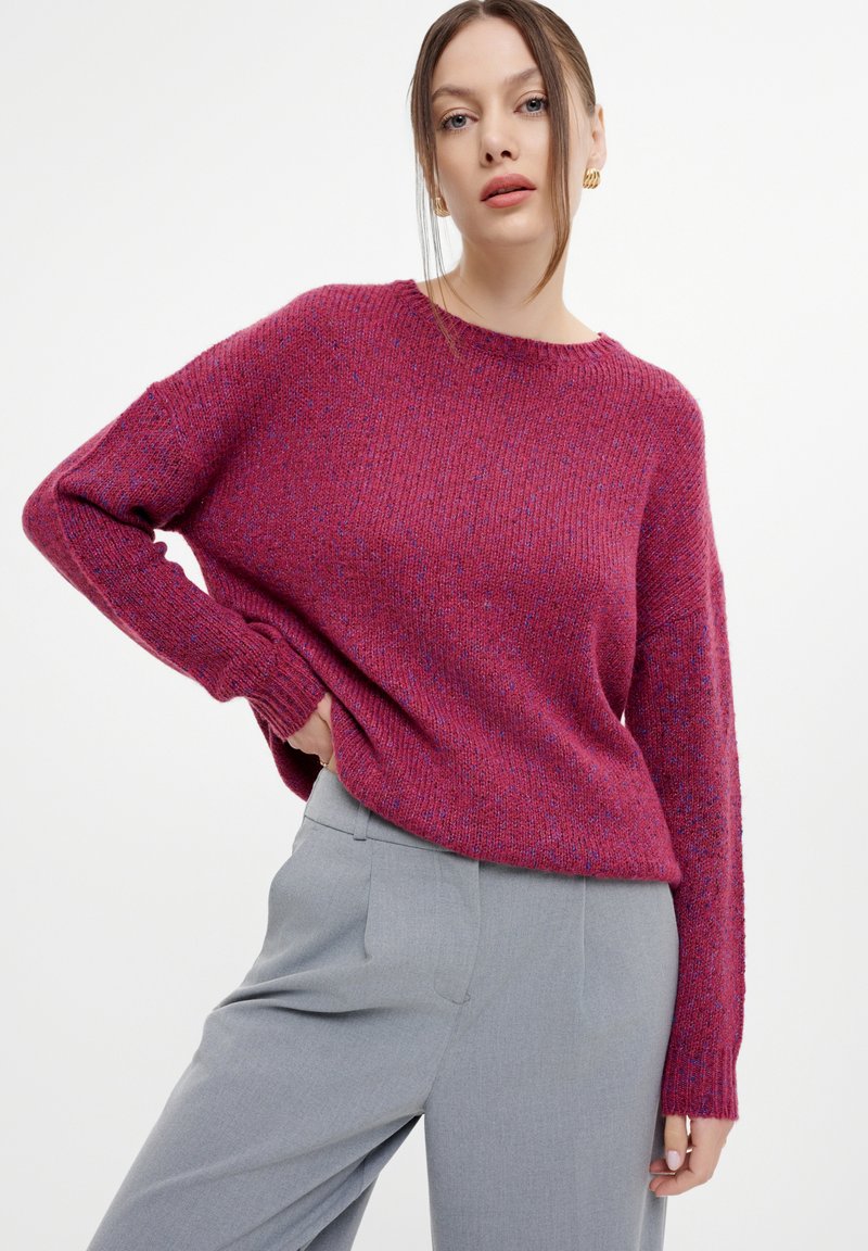 Maglione fucsia lavorato a maglia con maniche lunghe e scollatura rotonda, abbinato a pantaloni grigi chiari su misura. La trama appare morbida e accogliente.