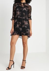 Robe noire à motif floral avec manches transparentes, ourlet à volants et détail d'encolure fendue. Associée à des talons hauts noirs à lanières.