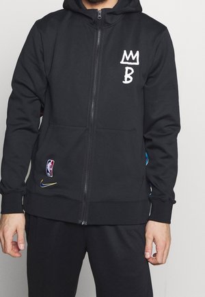 Man draagt een zwarte zip-up hoodie met wit "MB" en kroonsymbool op de borst, NBA- en Nike-logo's vlak bij de onderste zak.