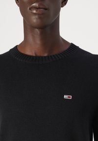 Svart strikkegenser med ribbestrikket halslinning. Har et lite brodert logo i rødt, hvitt og blått på brystet.