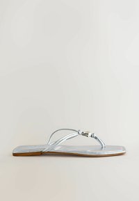 Sandale plate argentée avec des fines lanières et un petit détail de perle métallique sur un fond clair.