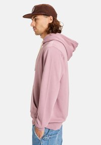 Sudadera rosa clara con cordón, bolsillo tipo canguro y puños acanalados, combinada con una gorra de pana marrón que presenta un parche de logo.