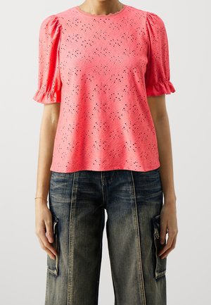 Różowy top z ażurowym wzorem, bufkami na rękawach i falbankowym dekoltem, z misternym wzorem wyciętym laserowo. Połączony z luźnymi dżinsami.