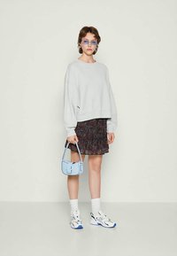 Jovem mulher vestindo um sweatshirt oversized cinza claro, saia floral, tênis brancos e óculos de sol azuis, segurando uma pequena bolsa azul clara.