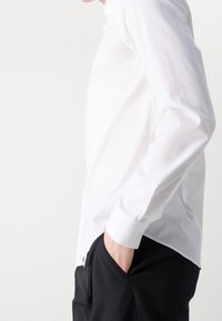 Persoon in een witte lange mouwen shirt en zwarte broek met één hand casual in de zak, van opzij weergegeven op een effen achtergrond.