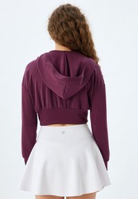 Sudadera corta morada con capucha y cintura recogida, combinada con una falda blanca de corte acampanado. Textura de tela suave, sin patrones ni acentos visibles.