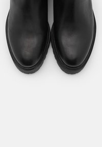 Mustat nahkaiset nilkkasaappaat, joissa on sileä pinta, pyöreä kärki ja paksu musta kumipohja. Minimalistinen muotoilu ilman näkyviä metalliosia.