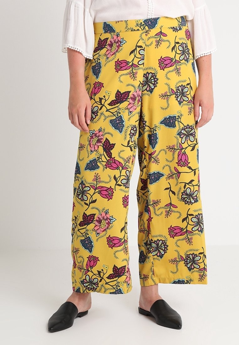 Pantalon jaune à jambes larges avec motif floral rose et bleu, porté avec une blouse blanche et des chaussures noires pointues sans lacets.
