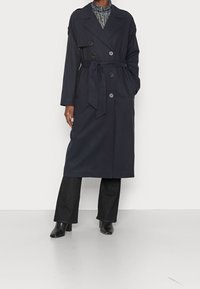 Marinblå trenchcoat med dubbelknäppta knappar, bälte i midjan, sidofickor och en slät, matta yta. Kombination med mörka jeans och klackar.