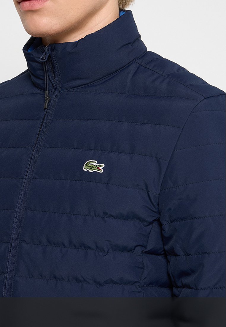 Veste matelassée zippée bleu marine avec col montant, présentant un design matelassé texturé et un logo de crocodile vert brodé sur la poitrine.