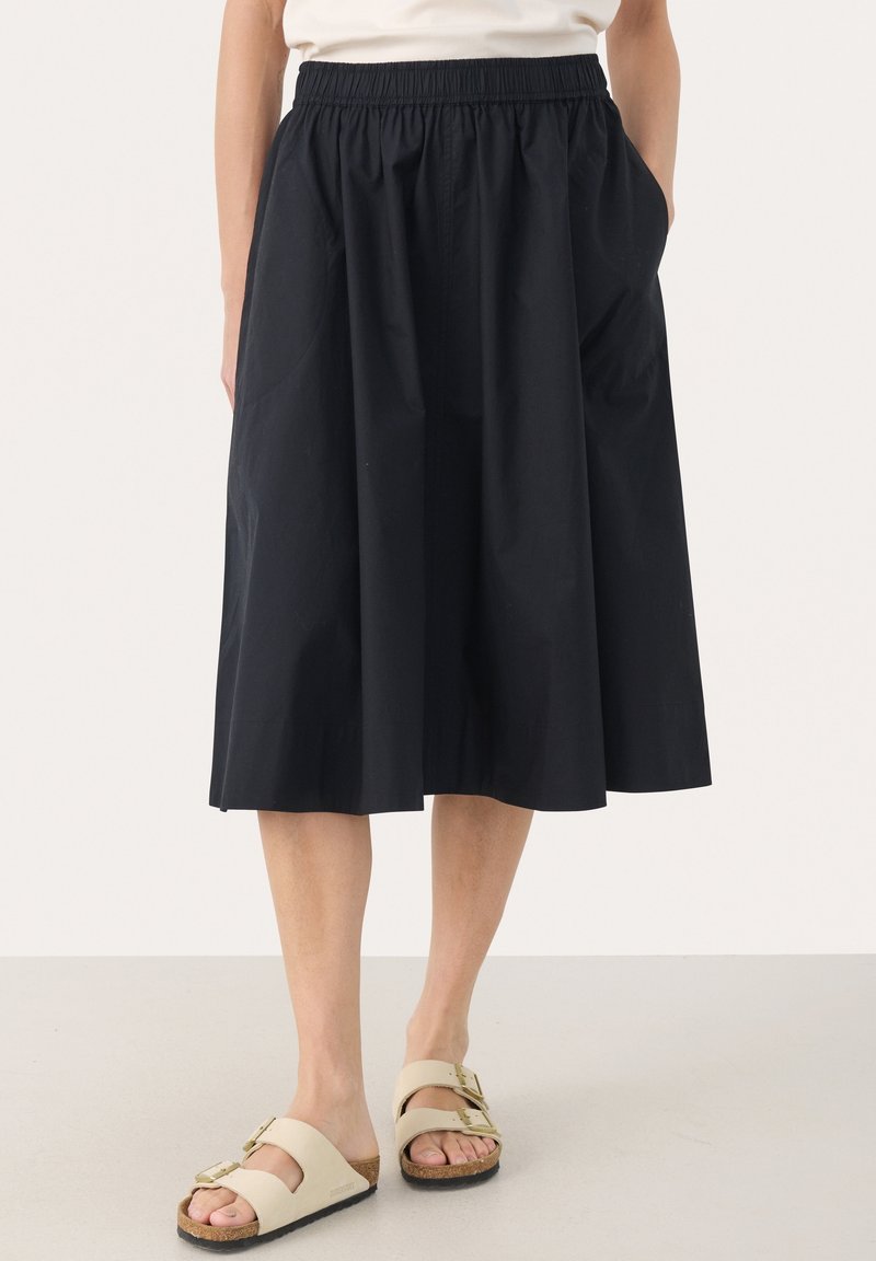 Personne portant une jupe noire longueur genou avec une taille élastique et des sandales beige à double bride, debout sur un sol uni.