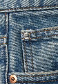 Tissu en denim bleu clair avec une texture de tissage visible, accentué par des éléments en cuivre. Présente des coutures orange contrastées et une petite poche.