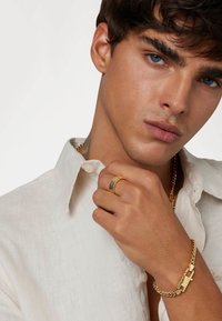 Bracelet en or avec un design texturé, bague en or et argent, et chemise en lin de couleur claire. Gros plan sur une main tenant le col de la chemise.