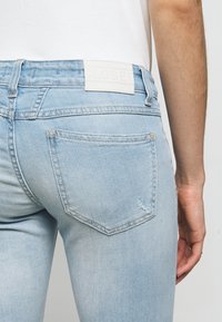 Jean en denim bleu clair avec une poche arrière, un léger effet délavé et une étiquette blanche sur la ceinture, porté avec une chemise blanche.