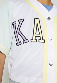 Maillot blanc à manches courtes avec des manches vert menthe, un liseré jaune, et les lettres "K" violettes avec un contour noir et "A" jaune. Il est orné de boutons-pression métalliques.