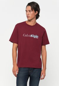 Young man wearing a burgundy Calvin Klein t-shirt and dark jeans standing against a plain white background, looking left.
