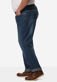 Jeans de mezclilla azul oscuro con corte recto, que cuentan con bolsillos laterales, bolsillos traseros y puños enrollados, combinados con zapatos marrones de cordones.